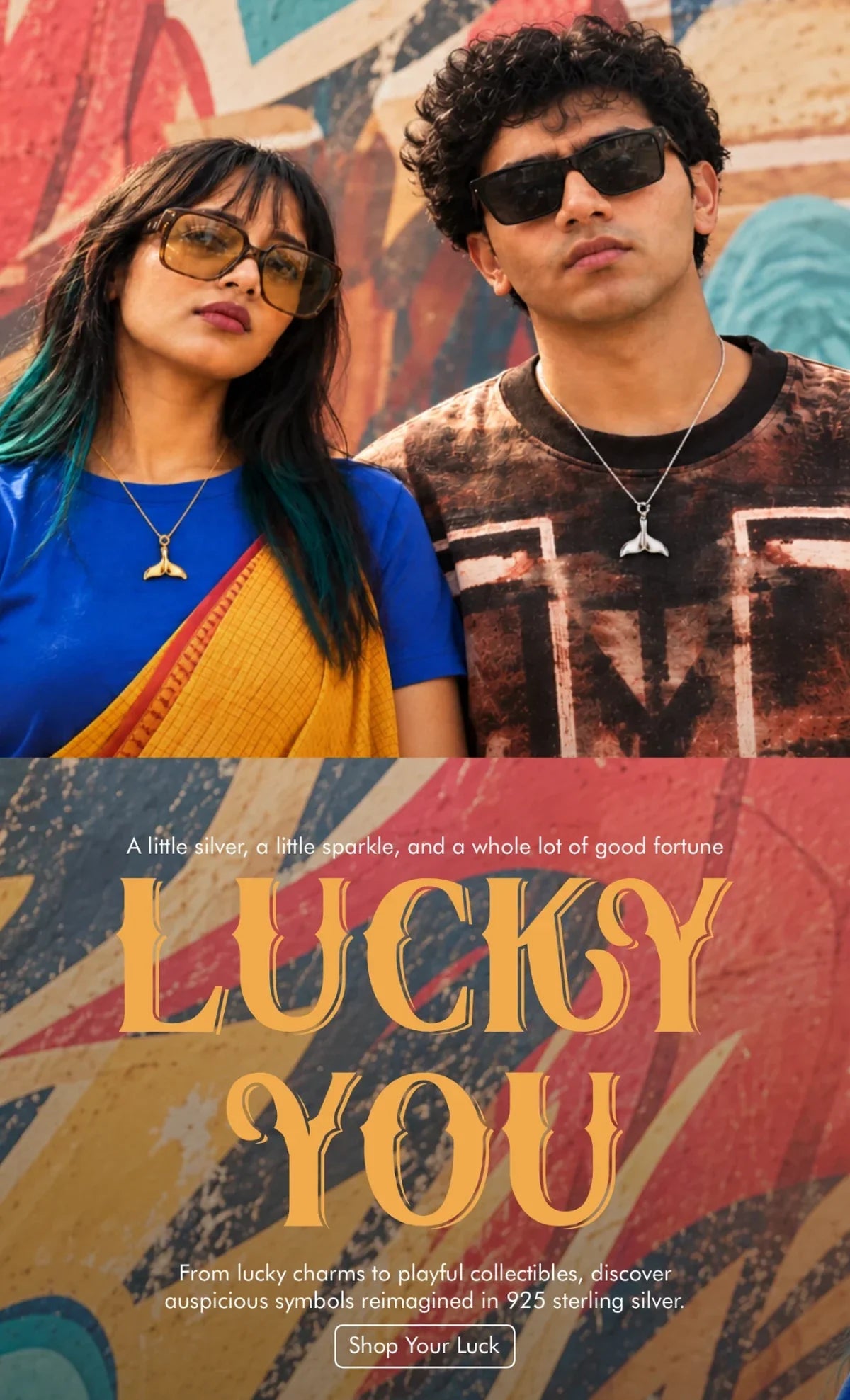 Lucky You collection: couple wearing silver and gold whale tail pendants against a painted wall
