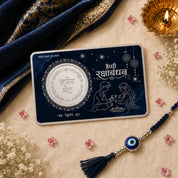 A silver Rakhi coin displayed on its dark blue presentation card, styled with a matching navy blue and silver evil eye Rakhi, a gold diya, and traditional Indian floral elements on a beige textured background.