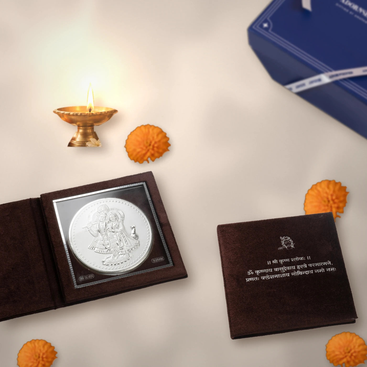 Blessings in Silver Radha Krishna edition: 999 silver Radha Krishna coin in its velvet shloka folder, with a brass diya, marigolds and the Adorn gift box